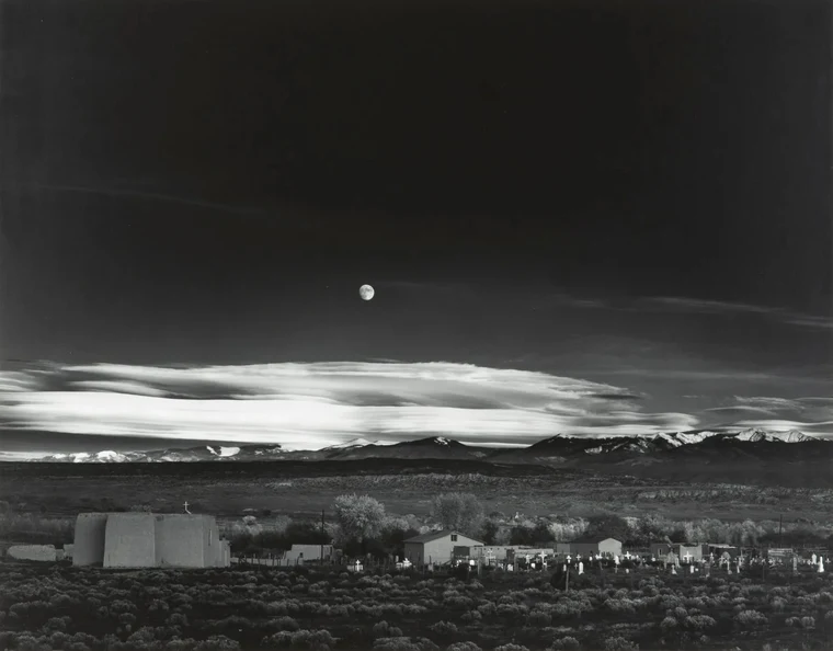 Ansel Adams Moonrise over Hernandez, a dark sky and moon above a luminous band of lit crosses and a dark foreground.