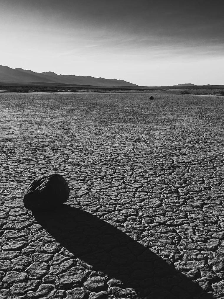 Black-and-white photograph of a single round dark rock on a cracked dry lake bed casting a long shadow toward the viewer, with distant mountains under a pale sky.