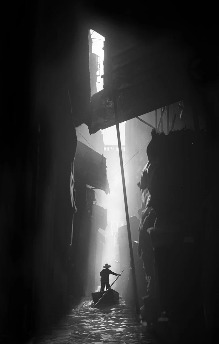 Black-and-white photograph of a small figure poling a boat through a narrow waterway, lit by a vertical shaft of light with shadowed walls and awnings closing in on both sides.