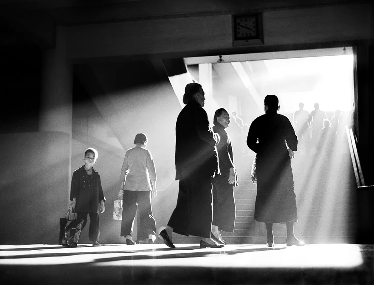 Black-and-white street photograph of backlit silhouetted figures standing in a shaft of light, strong shadow shapes around them.