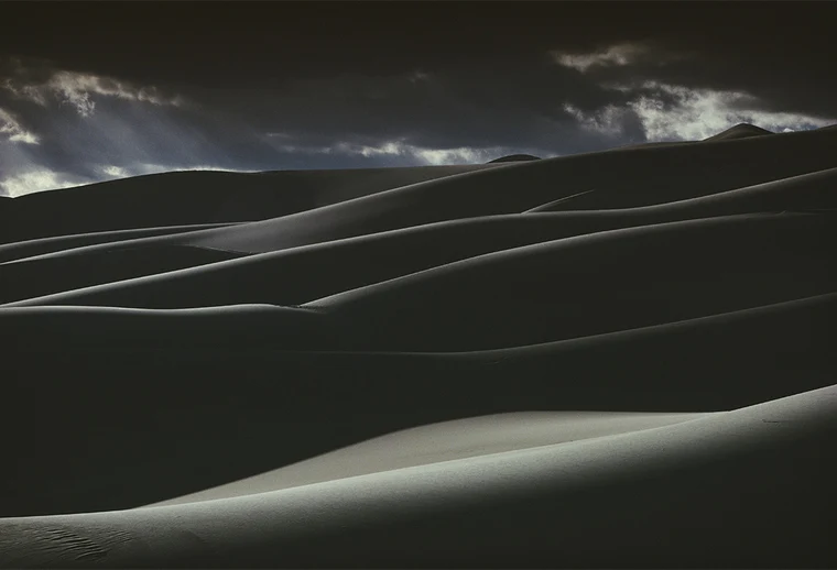Dark sand dunes lit only along their crests, falling in repeating curved ridges across the frame, with a narrow strip of cloudy blue sky at the top.