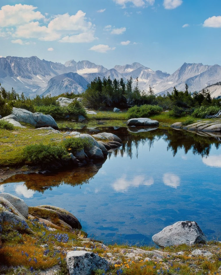 Color vertical photograph of a clear alpine lake reflecting jagged peaks and clouds, with granite boulders and wildflowers in the foreground.