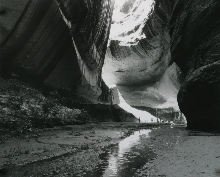 Black-and-white view inside a vast sandstone canyon, a wet channel leading the eye toward small silhouetted figures in the brightest passage.