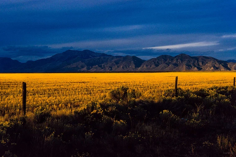 Golden sunlit plains stretching to dark distant mountains under a deep blue sky, with small fence posts in the foreground.