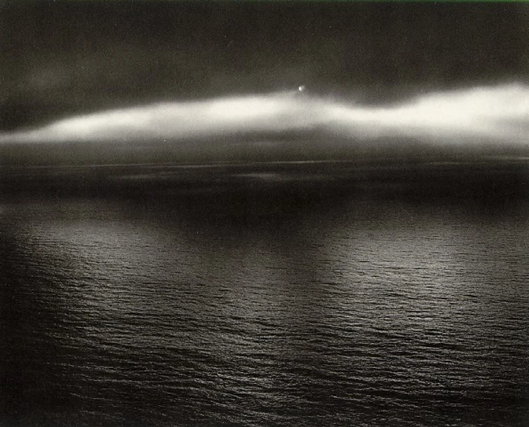 Dark moody seascape with a soft luminous cloud band hovering over a textured dark sea, a small bright point of light at the cloud's crest.
