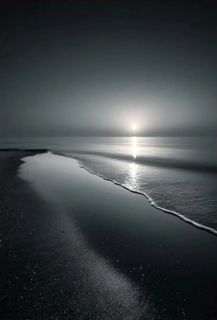 Black-and-white minimalist seascape with a small moon above a long stretch of beach, its reflection running down the wet sand.