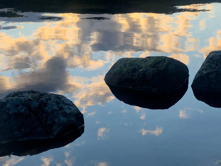 Three dark boulders in still water reflecting golden clouds, with a band of deep shadow at the top.