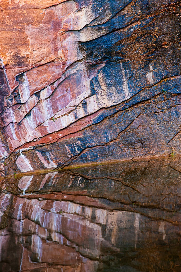 A reddish striated rock wall reflected in still water, the strata radiating in diagonal bands of color.