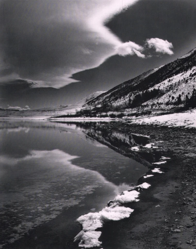 Black-and-white vertical landscape of a still lake, a large cloud form echoed by its reflection, ice along the shoreline, a dark ridge at right.