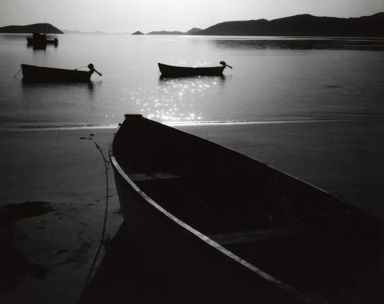 Dark silhouetted boats on a bay, heavy shadow in the foreground, scattered bright glints of light skimming the water.