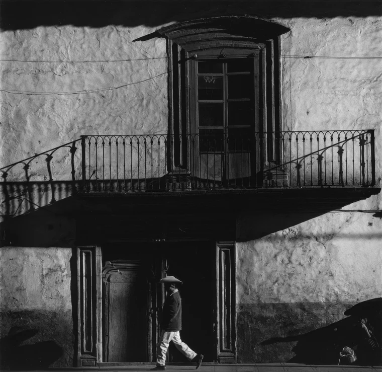 Architectural facade flattened into blocks of light and dark, a balcony reading as a pale trapezoid, a small figure crossing below.