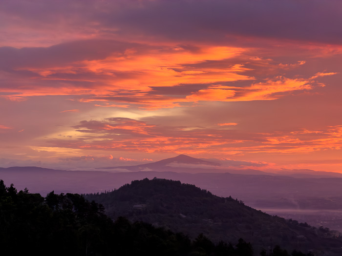 Glowing orange clouds above layered hills and a distant mountain at dawn